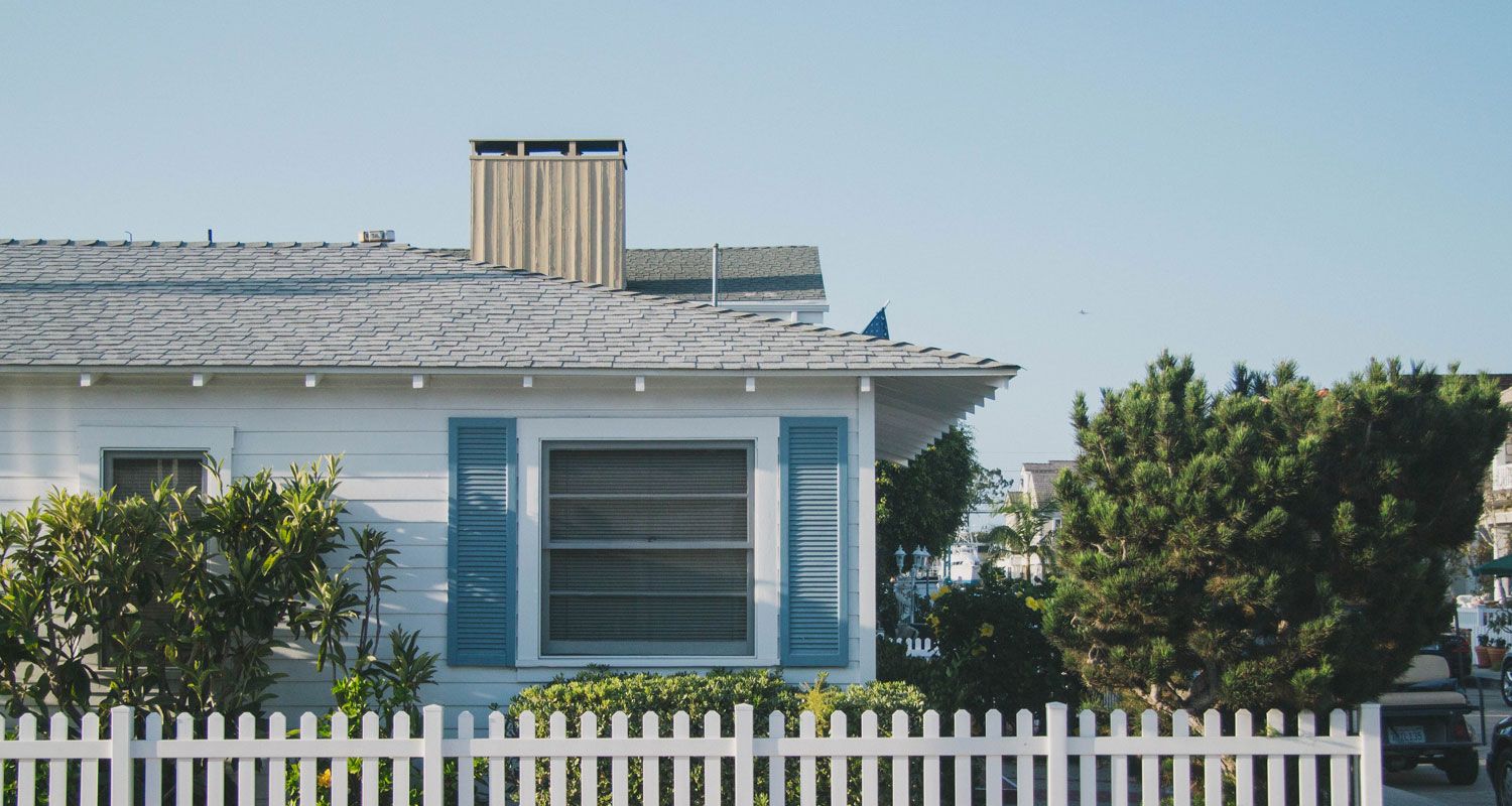 Window on white bungalow with blue exterior shutters sits behind some flora and a white picket fence