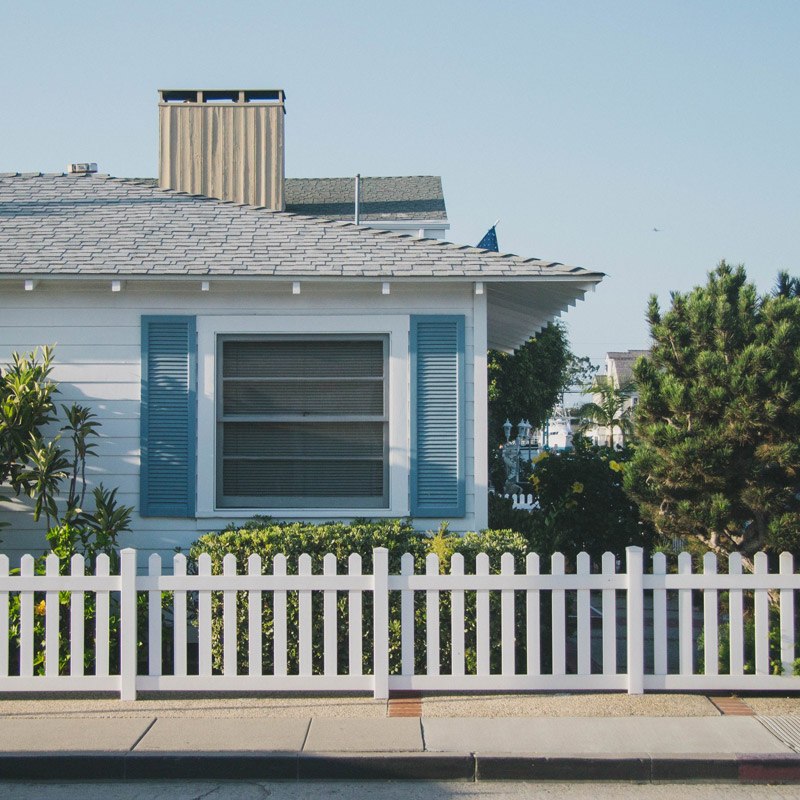 Window on white bungalow with blue exterior shutters sits behind some flora and a white picket fence