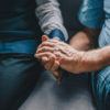 An elderly man wearing a gold wedding band holds another person's hand with both hands